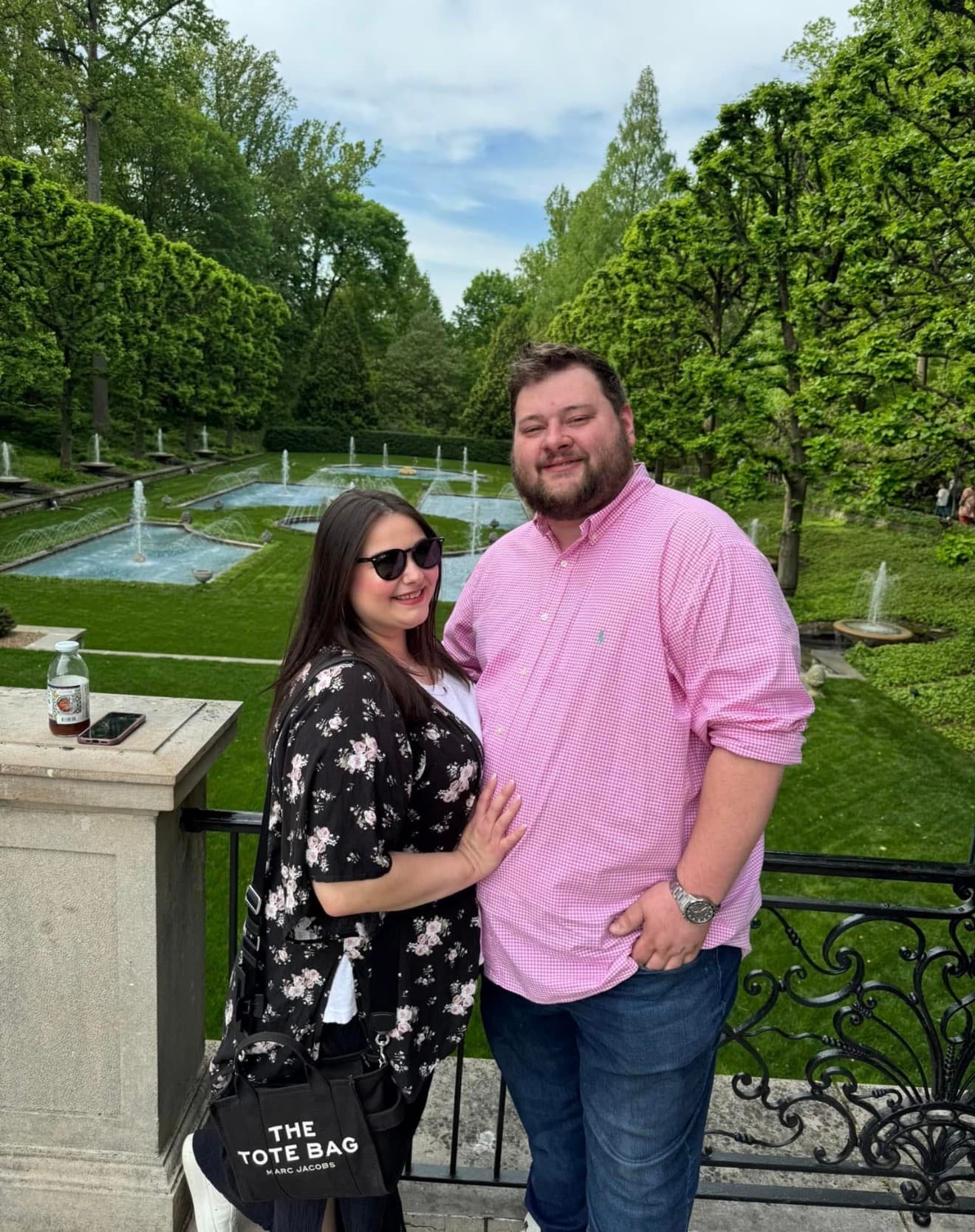Smiling couple on a balcony overlooking a formal garden with multiple fountains.